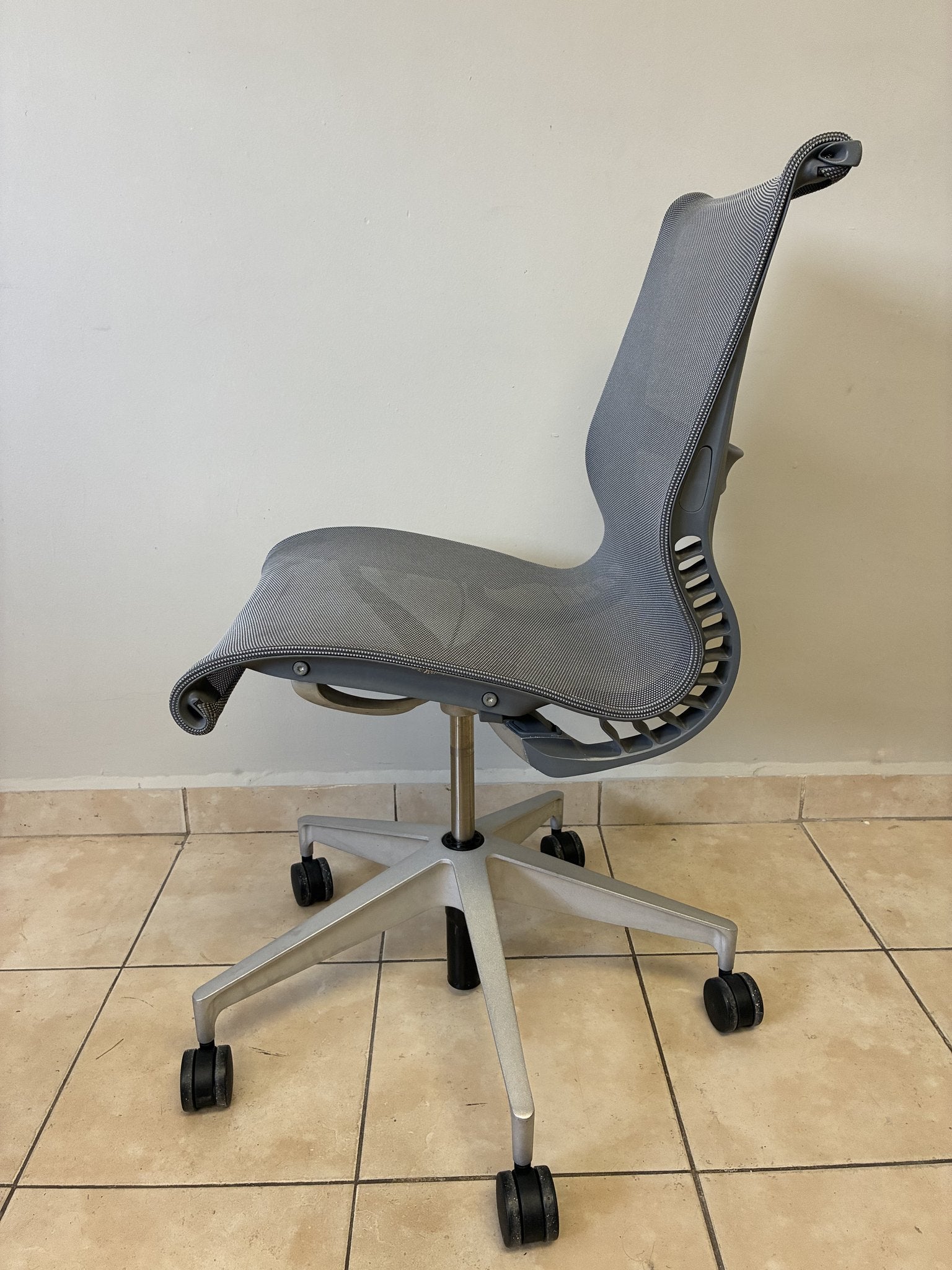 Silla de oficina Herman Miller Setu preowned, sin brazos, en color gris claro, vista lateral sobre piso de azulejos.