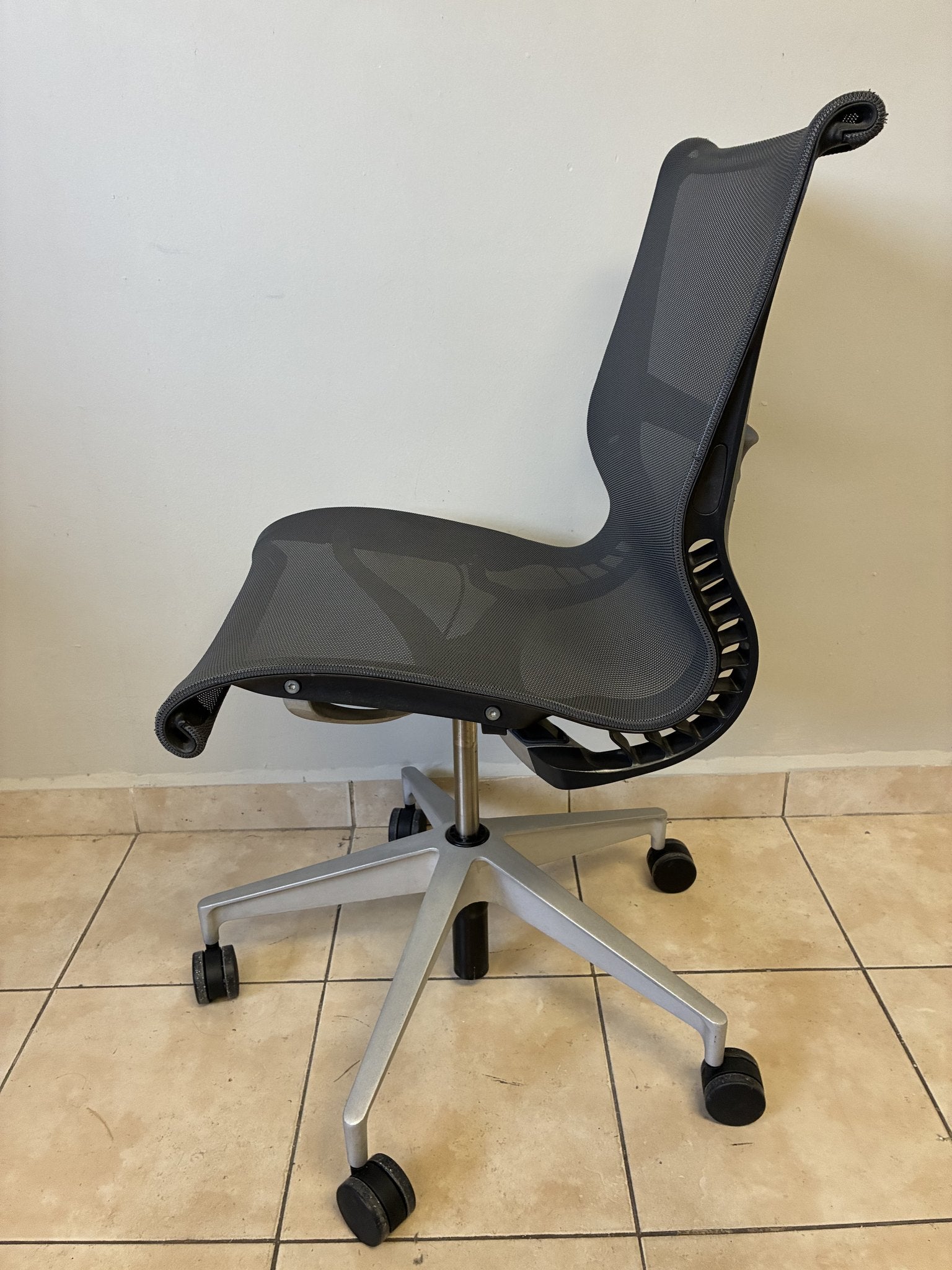 Silla de oficina Herman Miller Setu pre-owned, sin brazos, en gris oscuro, vista lateral sobre piso de azulejos.