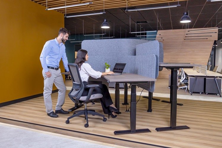 Dos personas en una estación de trabajo con escritorio de altura ajustable y silla ergonómica de malla